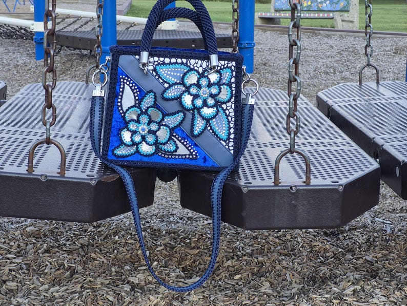 May include: A square-shaped navy blue handbag featuring a decorative floral pattern with silver and light blue accents. The bag includes a braided navy blue handle, a matching shoulder strap, and silver-toned hardware attachments on the sides.