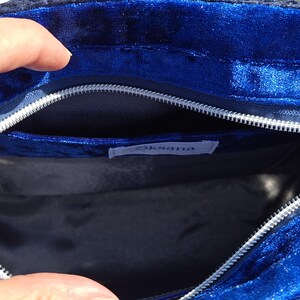 May include: A deep blue velvet bag interior featuring a silver zipper closure along the top edge. The inside is lined with a dark fabric and includes a small white rectangular label with the text Oksana stitched onto the inner pocket area.