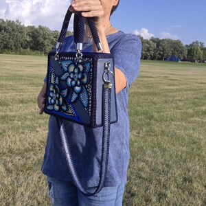 May include: A structured rectangular handbag in shades of blue and grey, featuring a decorative floral pattern with intricate dot-work detailing. The bag includes two braided top handles, silver-toned hardware, and a detachable matching shoulder strap.