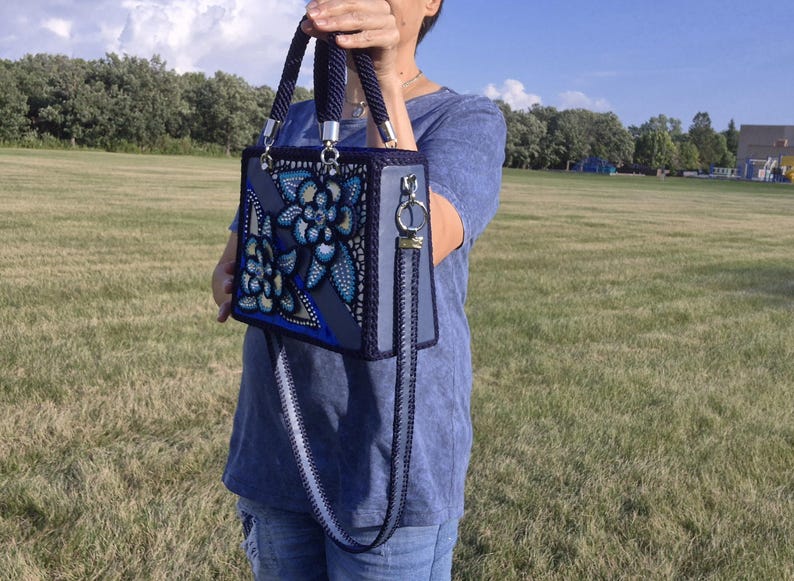 May include: A structured rectangular handbag in shades of blue and grey, featuring a decorative floral pattern with intricate dot-work detailing. The bag includes two braided top handles, silver-toned hardware, and a detachable matching shoulder strap.