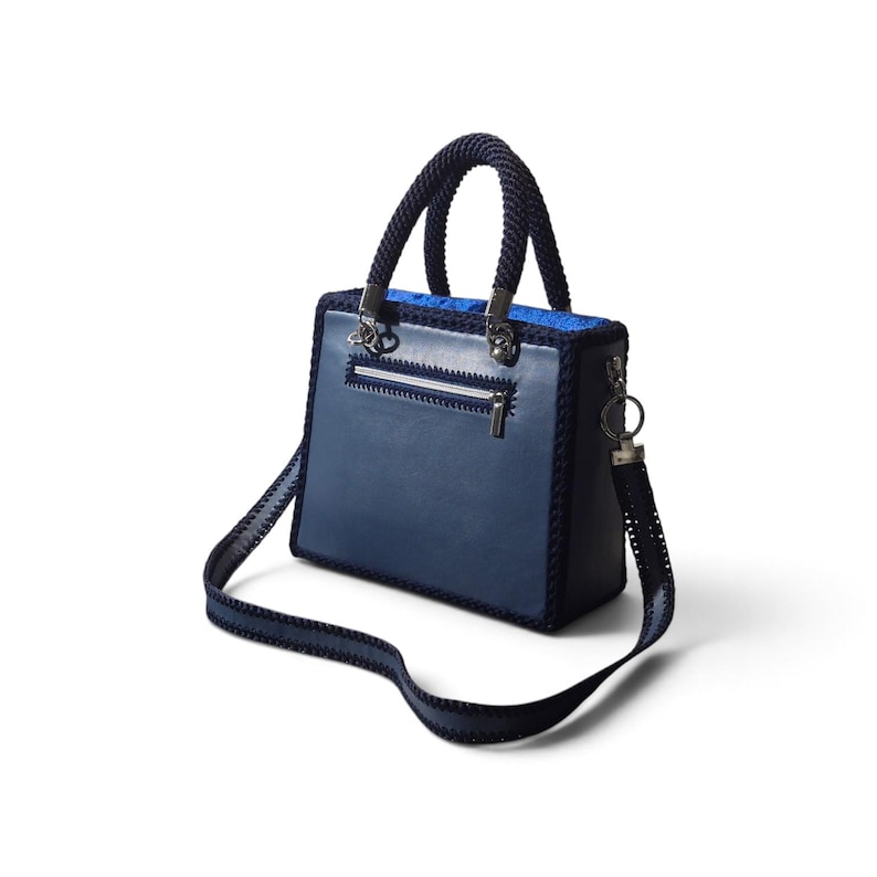 May include: A structured navy blue handbag featuring a smooth front panel, a zippered pocket, and dark blue braided rope handles. The bag includes a matching detachable shoulder strap with stitched edges and silver-toned metal hardware accents.