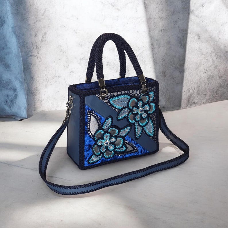 May include: A structured handbag in shades of blue, featuring a dark blue braided handle and shoulder strap. The exterior displays a blue velvet panel with intricate floral lace overlays, accented by light blue and silver sequin embroidery.