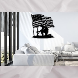 May include: A black metal wall art piece depicting a kneeling soldier in silhouette, with a waving American flag and a helmet and rifle. The artwork is mounted on a white wall above a white sofa, with a window and blinds in the background.