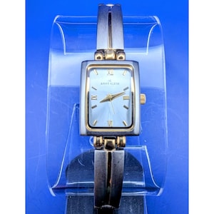 May include: Anne Klein rectangular wristwatch with a silver and gold-tone metal band and case. The watch face is light blue with gold hands and Roman numeral hour markers. The watch is displayed on a clear plastic stand.