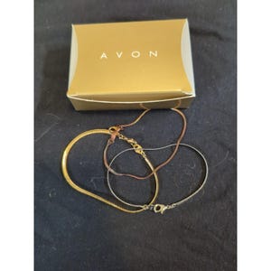 May include: A gold-colored necklace with a snake chain design, a copper-colored necklace, and a silver-colored necklace are displayed next to a gold-colored box with the word "AVON" printed on it. The necklaces are on a dark blue surface.