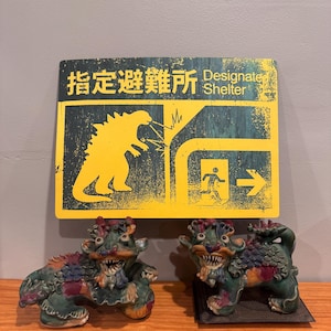 May include: Two ceramic guardian lion statues with green, blue, and red details, flanking a yellow sign with Japanese text and the words "Designate Shelter." The sign features a Godzilla-like figure and an arrow.