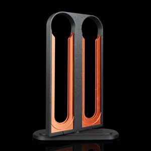 Peut inclure: Un support vertical noir à deux fentes avec une finition extérieure texturée et des canaux intérieurs lisses de couleur cuivre métallique. La structure repose sur une base noire plate de forme ovale, conçue pour maintenir deux objets à la verticale.