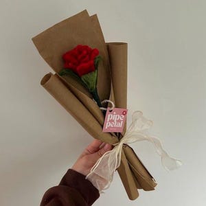 May include: A single red rose with green leaves, presented as a bouquet wrapped in brown paper and tied with a cream ribbon. The bouquet has a pink tag with the text "pipe petal".