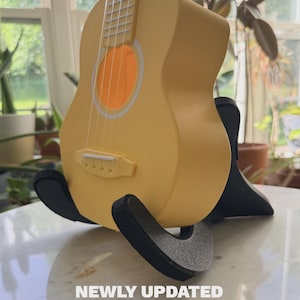 May include: A yellow ukulele rests on a black stand. The ukulele has a round sound hole with an orange interior and white trim. The text "NEWLY UPDATED" and "NOW WITH 3MM THICK FELT TO PROTECT YOUR UKE FINISH" is visible at the bottom of the image.