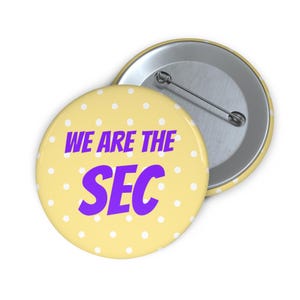 May include: A round pin-back button featuring a pale yellow background with a white polka dot pattern. The text WE ARE THE SEC is printed in bold purple capital letters across the center. The back shows a silver metal pin mechanism for attachment.