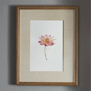 May include: A framed watercolor painting featuring a single pink lotus flower with a yellow center on a white background. The artwork is encased in a rectangular gold frame with a beaded edge and a textured beige matting surrounding the print.