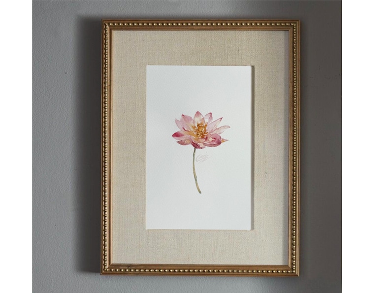 May include: A framed watercolor painting featuring a single pink lotus flower with a yellow center on a white background. The artwork is encased in a rectangular gold frame with a beaded edge and a textured beige matting surrounding the print.
