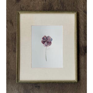 May include: A framed watercolor painting on white paper featuring a single purple and blue flower with a thin green stem. The artwork is enclosed in a light-colored fabric mat and a thin, textured, muted green wooden frame with a square profile.