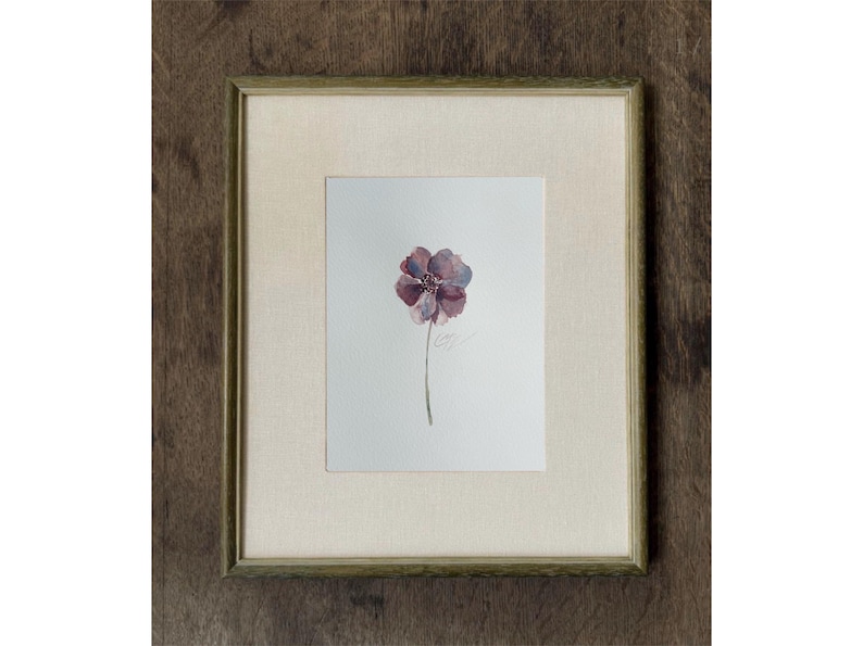 May include: A framed watercolor painting on white paper featuring a single purple and blue flower with a thin green stem. The artwork is enclosed in a light-colored fabric mat and a thin, textured, muted green wooden frame with a square profile.