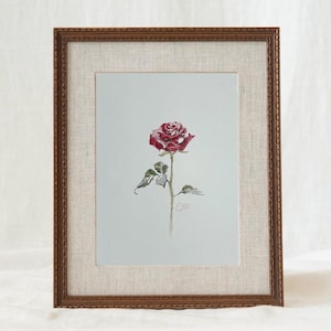 May include: A framed watercolor painting of a single red rose with green leaves on a light gray background. The artwork is enclosed in a textured beige mat and a dark brown wooden frame with a decorative, ridged edge and a subtle pattern.