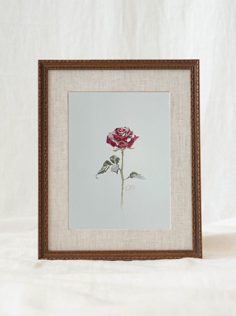 May include: A framed watercolor painting of a single red rose with green leaves on a light gray background. The artwork is enclosed in a textured beige mat and a dark brown wooden frame with a decorative, ridged edge and a subtle pattern.