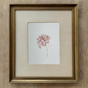 May include: A watercolor painting of a single pink flower with delicate petals and a thin green stem, centered on a white background. The artwork is encased in a rectangular gold-toned frame with a textured, off-white fabric matting border.