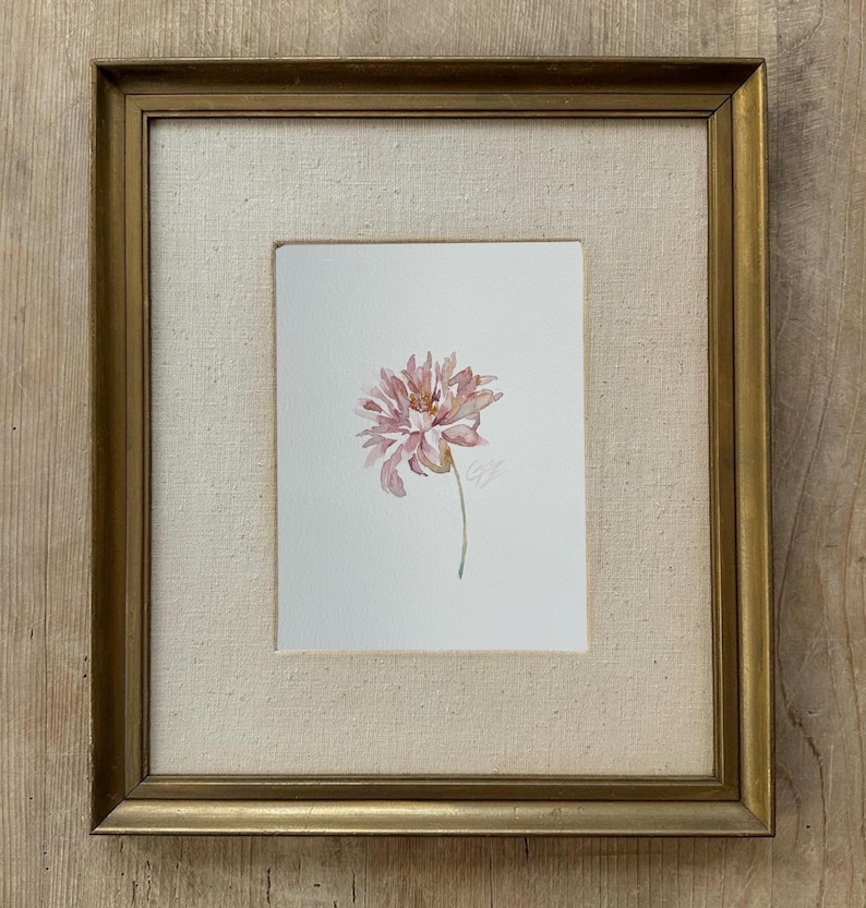 May include: A watercolor painting of a single pink flower with delicate petals and a thin green stem, centered on a white background. The artwork is encased in a rectangular gold-toned frame with a textured, off-white fabric matting border.