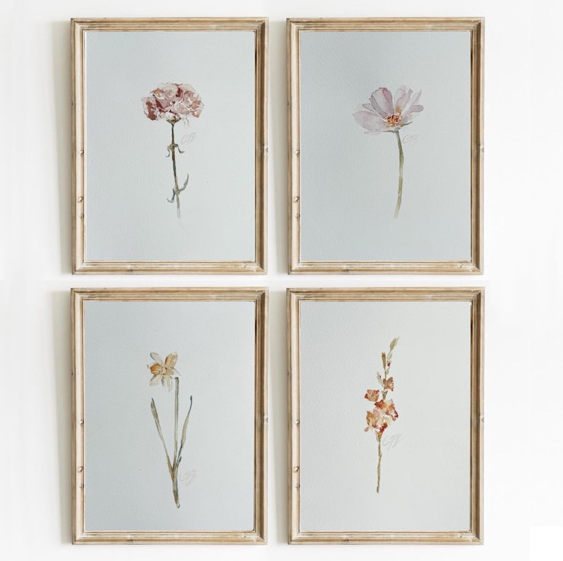 May include: A set of four rectangular wall art prints in light wood frames, each featuring a single watercolor botanical illustration on a pale gray background. The prints depict a pink carnation, a light purple cosmos, a yellow daffodil, and orange flowers.