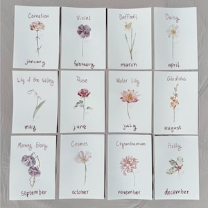 May include: A set of twelve rectangular cards featuring watercolor illustrations of birth month flowers. Each card displays a flower name and the corresponding month in handwritten text, including Carnation, Violet, Daffodil, Daisy, Lily of the Valley, Rose, Water Lily, Gladiolus, Morning Glory, Cosmos, Chrysanthemum, and Holly.