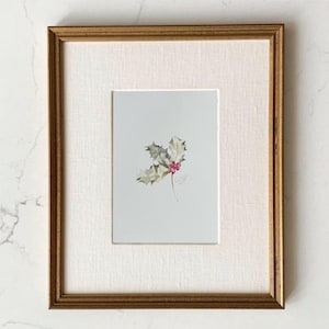May include: A framed watercolor painting featuring a small sprig of green holly leaves with bright red berries centered on a light gray background. The artwork is encased in a textured off-white mat and a thin, metallic gold-toned wooden frame.