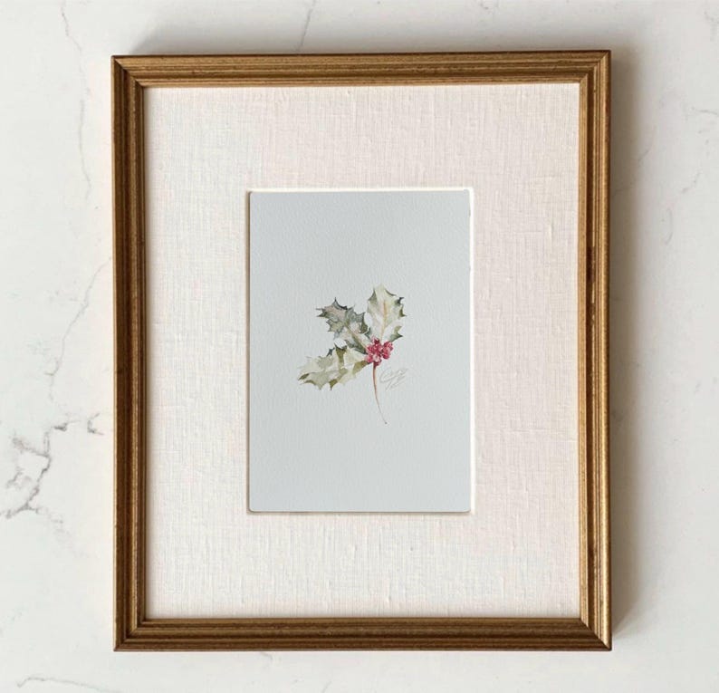 May include: A framed watercolor painting featuring a small sprig of green holly leaves with bright red berries centered on a light gray background. The artwork is encased in a textured off-white mat and a thin, metallic gold-toned wooden frame.