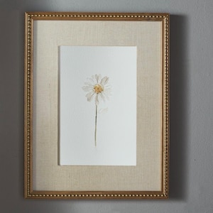 May include: A framed watercolor painting of a single white daisy with a yellow center on a white background. The artwork is encased in a rectangular gold frame with a beaded edge and a textured beige fabric mat surrounding the central image.