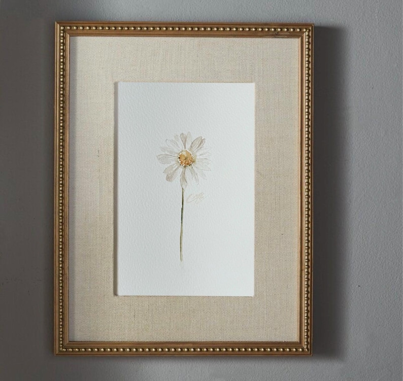 May include: A framed watercolor painting of a single white daisy with a yellow center on a white background. The artwork is encased in a rectangular gold frame with a beaded edge and a textured beige fabric mat surrounding the central image.
