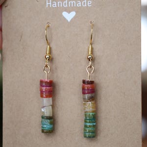 Peut inclure: Boucles d'oreilles faites à la main avec des crochets dorés. Chaque boucle d'oreille présente une pile de perles plates colorées dans les tons de rouge, orange, blanc, vert et bleu. Les boucles d'oreilles sont présentées sur une carte marron avec le mot "Handmade" et un cœur.