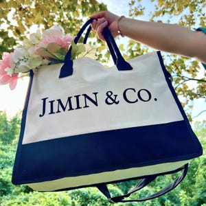 May include: A cream-colored tote bag with a navy blue bottom panel and handles. The text "JIMIN & CO." is printed in black on the front. The bag is filled with pink and white flowers. The bag is held up against a background of green foliage.