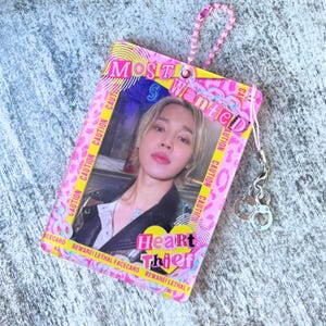 Puede incluir: Una tarjeta rosa con las palabras "MOST WANTED" y "Heart Thief". Presenta una foto de una persona con cabello rubio. Una cadena de cuentas rosas y un dije de esposas están adjuntos.