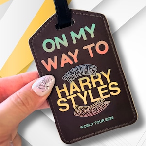 May include: A dark brown rectangular luggage tag with a black strap. The text reads ON MY WAY TO TOGETHER HARRY STYLES TOGETHER WORLD TOUR 2026. The words ON MY WAY TO are in a gradient, HARRY STYLES is in yellow, and WORLD TOUR 2026 is in light blue.