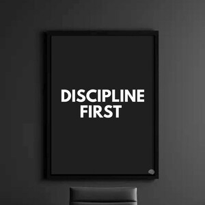May include: A framed black and white print with the words "DISCIPLINE FIRST" in bold, white capital letters. The print is in a black frame and hangs on a dark gray wall. A black chair is visible below the frame.