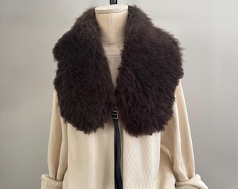 May include: A cream-colored ribbed knit sweater featuring a large, dark brown faux fur collar. The collar is secured at the center front with a thin black leather strap and a small silver-toned metal buckle. The sweater has long sleeves.