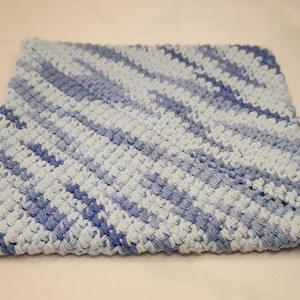 May include: A square, hand-crocheted washcloth in shades of light blue and white. The diagonal pattern creates a textured appearance. The washcloth is made of soft yarn.