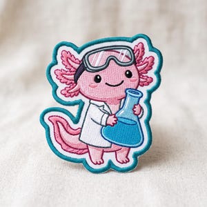May include: An embroidered patch featuring a pink axolotl wearing a white lab coat and grey safety goggles. The axolotl is holding a blue conical flask filled with blue liquid. The patch has a thick teal border and a white outline.