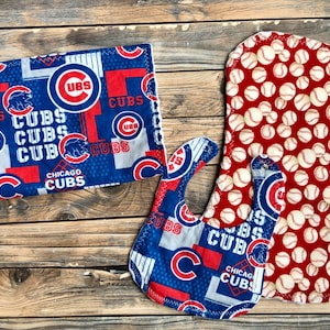 May include: A set of Chicago Cubs themed baby items, including a bib, burp cloth, and a rectangular item. The bib and rectangular item feature the Cubs logo and text on a blue background. The burp cloth has a red background with baseballs.