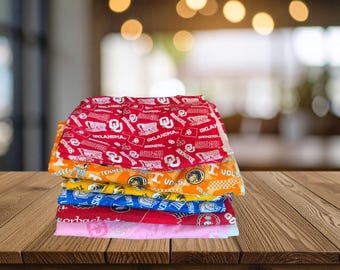 May include: A stack of folded fabric pieces featuring various collegiate sports team logos and colors. The top layer is red with white Oklahoma Sooners branding, followed by orange Tennessee Volunteers, yellow, blue, red, and light pink patterned fabrics.