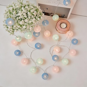 Pode incluir: Uma guirlanda de luzes pastel em forma de coração, com bolas rosa, azuis e verdes. As luzes estão em um fio de prata fino. Um pequeno castiçal e um buquê de flores brancas no fundo.