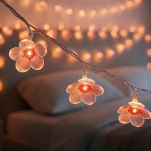 May include: A string of flower-shaped fairy lights with a warm, orange glow. The lights are strung on a silver wire and are arranged in a bedroom setting. The flower lights are made of a translucent material.