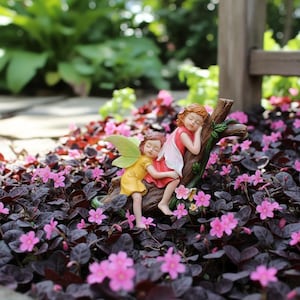 May include: A fairy figurine set featuring two sleeping fairies. One fairy wears a yellow dress with green wings, the other wears a red dress with white wings. They are resting on a brown tree branch, surrounded by pink flowers.