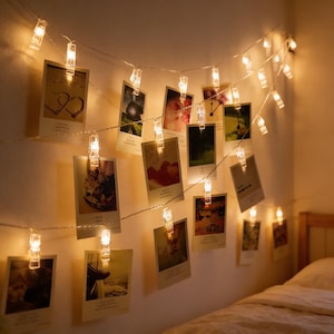 May include: A string of warm white LED lights with clear photo clips, displaying several small photographs. The lights are strung across a wall, creating a cozy and decorative ambiance. The photos are of various sizes and feature different images.