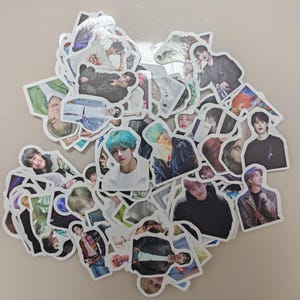 May include: A pile of die-cut stickers, each displaying a different image of a person. The stickers have white borders and are scattered on a light surface. The images show a variety of hairstyles and clothing styles. The stickers are of various sizes.