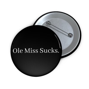 May include: Two round black buttons with a metal pin backing. The front button has the text "Ole Miss Sucks." in white, centered on the black surface. One button is face up, and the other is slightly angled.