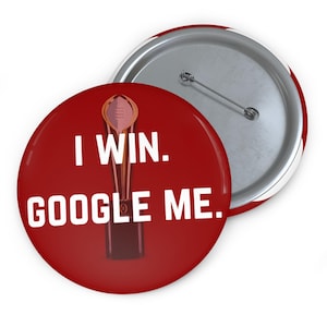 May include: Red button with the text "I WIN. GOOGLE ME." in white. The button features a graphic of a trophy. The back of the button is silver with a pin.