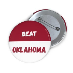 May include: A round button with a red and white design. The top half is red with the word "BEAT" in white. The bottom half is white with the word "OKLAHOMA" in red. A second button is visible in the background.