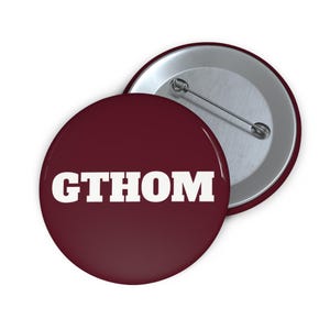 May include: Two round maroon buttons with the white text "GTHOM". One button is face up, displaying the text, and the other is face down, showing the pin back. The buttons are likely for clothing or accessories.