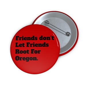 May include: A red button with the text "Friends don't Let Friends Root For Oregon." The button has a silver pin back and a shiny, glossy finish. The button is round and has a metallic silver edge.
