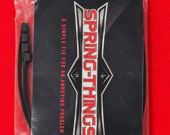 Puede incluir: Un paquete de producto rectangular negro dentro de una bolsa de plástico transparente. El paquete presenta el texto blanco y rojo SPRING-THINGS y la frase A SIMPLE FIX FOR AN ANNOYING PROBLEM. Varios cordones negros finos y flexibles son visibles.