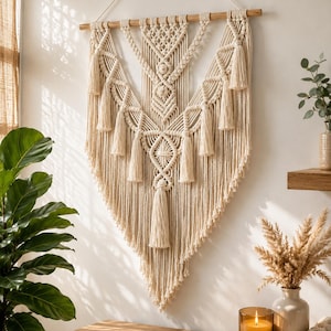 May include: A large off-white macrame wall hanging suspended from a horizontal wooden dowel. The piece features intricate knotted patterns, including diamond shapes and central vertical columns, finished with multiple hanging tassels along the edges.